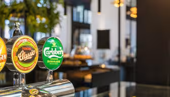 Nærbilde av ølkraner med merkevarene Tuborg Classic og Carlsberg i moderne barmiljø. Bakgrunnen er mykt uskarp med glimt av innendørs planter og omgivelseslys.