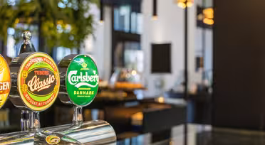 Nærbilde av ølkraner med merkevarene Tuborg Classic og Carlsberg i moderne barmiljø. Bakgrunnen er mykt uskarp med glimt av innendørs planter og omgivelseslys.