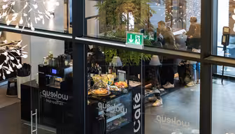 Utsikt gjennom glassvegger inn i en moderne kafé med folk som sitter og står. Dekorasjoner og et nødutgangsskilt er synlige. En disk viser bakverk og en kaffemaskin.
