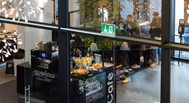 Utsikt gjennom glassvegger inn i en moderne kafé med folk som sitter og står. Dekorasjoner og et nødutgangsskilt er synlige. En disk viser bakverk og en kaffemaskin.