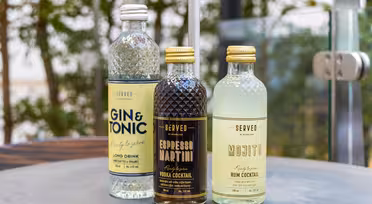 Tre flaskecocktailer vises på et grått bord utendørs: en klar flaske «Gin & Tonic», en mørk flaske «Espresso Martini» og en lysegrønn flaske «Mojito». Uskarpt grønt er synlig i bakgrunnen.