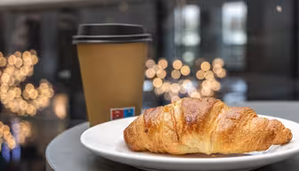 En gyllenbrun croissant sitter på en hvit tallerken ved siden av en brun takeaway-kaffekopp med svart lokk. Bakgrunnen er mykt uskarp, med sirkulære bokeh-lys som skaper en koselig atmosfære.