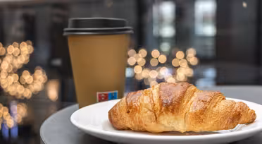 En gyllenbrun croissant sitter på en hvit tallerken ved siden av en brun takeaway-kaffekopp med svart lokk. Bakgrunnen er mykt uskarp, med sirkulære bokeh-lys som skaper en koselig atmosfære.
