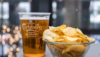 En klar plastkopp fylt med Carlsberg-øl settes ved siden av en glassbolle med potetgull på et bord. Bakgrunnen har uskarpt lys, noe som gir en koselig atmosfære til scenen.