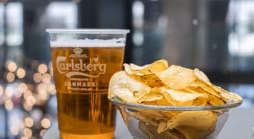En klar plastkopp fylt med Carlsberg-øl settes ved siden av en glassbolle med potetgull på et bord. Bakgrunnen har uskarpt lys, noe som gir en koselig atmosfære til scenen.