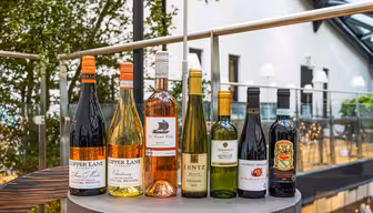 Et utvalg av åtte vinflasker, inkludert røde, hvite og en rosé, arrangert på et rundt bord. Bakgrunnen har et lyst rom med grøntområder og store vinduer, som skaper en innbydende atmosfære.