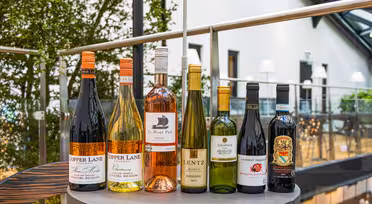 Et utvalg av åtte vinflasker, inkludert røde, hvite og en rosé, arrangert på et rundt bord. Bakgrunnen har et lyst rom med grøntområder og store vinduer, som skaper en innbydende atmosfære.