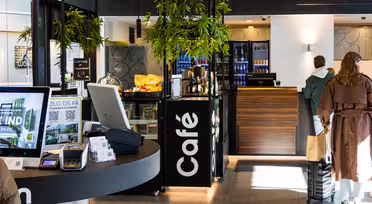 Moderne kaféinteriør med digitale menyer, kaffedisk og noen få bestillere. Plassen er dekorert med grønne planter og har en moderne design. Betalingsterminaler er synlige på en sirkulær skranke. Naturlig lys lyser opp området.