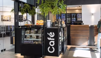 Moderne kaféinteriør med en svart disk som viser snacks og en kaffemeny. Hyller med produkter er synlige, sammen med en resepsjonsskranke i tre i bakgrunnen. Overhead er det grønne hengende planter. Sollyset strømmer gjennom inngangen.