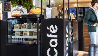 Et moderne kaféinteriør med en svart disk som viser ulike snacks, kaffe og vinflasker. En mann i grønn jakke med bagasje står i nærheten. Grønne planter henger fra taket, og legger til et snev av natur til rommet.