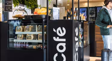 Et moderne kaféinteriør med en svart disk som viser ulike snacks, kaffe og vinflasker. En mann i grønn jakke med bagasje står i nærheten. Grønne planter henger fra taket, og legger til et snev av natur til rommet.