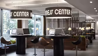 Moderne forretningssenter med elegant innredning, med runde bord med datamaskiner, omgitt av stoler. Store trommelys henger over, og en treskillevegg skiller området. Naturlig lys strømmer inn fra store vinduer, og forbedrer den moderne atmosfæren.