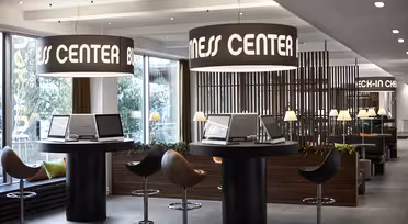 Moderne forretningssenter med elegant innredning, med runde bord med datamaskiner, omgitt av stoler. Store trommelys henger over, og en treskillevegg skiller området. Naturlig lys strømmer inn fra store vinduer, og forbedrer den moderne atmosfæren.