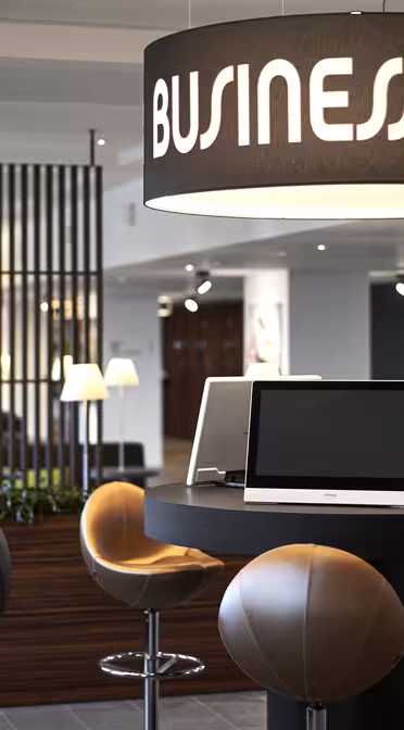 Moderne forretningssenterinteriør med elegante svarte bord og barkrakker. Store runde lysarmaturer med «Business Center»-tekst henger i taket. Datamaskiner er plassert på bordene, og rommet har et tidsriktig, profesjonelt design.
