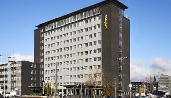 En moderne hotellbygning i flere etasjer med en elegant svart fasade og store vinduer. Hotellets navn vises vertikalt i øvre hjørne. Forgrunnen har biler, trær og en klar blå himmel.