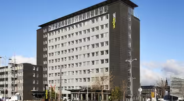 En moderne hotellbygning i flere etasjer med en elegant svart fasade og store vinduer. Hotellets navn vises vertikalt i øvre hjørne. Forgrunnen har biler, trær og en klar blå himmel.