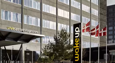 Moderne hotellbygg med store vinduer og skiltet "Wakeup Copenhagen" synlig. Tre danske flagg vaier på stenger utenfor inngangen. Noen få små trær er foran bygningen.