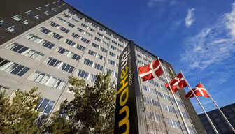 En høy bygning med et skilt der det står "Wakeup Copenhagen" står mot en klar blå himmel. Tre danske flagg vaier på flaggstenger foran bygningen, omgitt av noen trær.