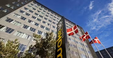En høy bygning med et skilt der det står "Wakeup Copenhagen" står mot en klar blå himmel. Tre danske flagg vaier på flaggstenger foran bygningen, omgitt av noen trær.