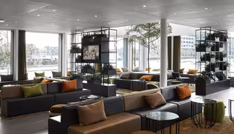 Moderne hotellobby med store vinduer, mørke sofaer med oransje og beige puter. Grønne planter står utstilt i hyller, og runde glassbord er spredt utover. Naturlig lys lyser opp rommet og skaper en innbydende atmosfære.