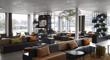 Moderne hotellobby med store vinduer, mørke sofaer med oransje og beige puter. Grønne planter står utstilt i hyller, og runde glassbord er spredt utover. Naturlig lys lyser opp rommet og skaper en innbydende atmosfære.