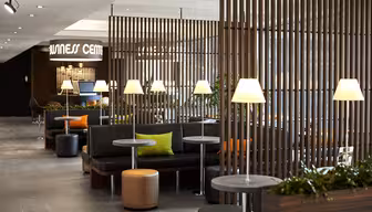 Moderne salong med svarte sitteplasser, oransje og grønne puter, runde bord og tente lamper. Trelameller deler opp rommet, og planter dekorerer området. Et skilt for et "Business Center" er i bakgrunnen.