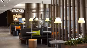 Moderne salong med svarte sitteplasser, oransje og grønne puter, runde bord og tente lamper. Trelameller deler opp rommet, og planter dekorerer området. Et skilt for et "Business Center" er i bakgrunnen.