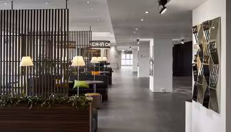 Moderne hotellobby med elegant design, med treskillevegger, koselige sitteplasser med grønne og oransje puter og moderne belysning. En geometrisk veggskulptur pryder høyre side, og gir et stilig preg.