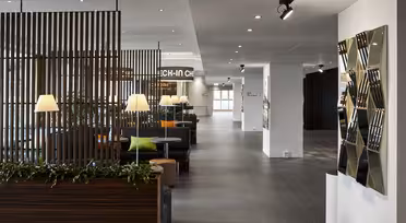 Moderne hotellobby med elegant design, med treskillevegger, koselige sitteplasser med grønne og oransje puter og moderne belysning. En geometrisk veggskulptur pryder høyre side, og gir et stilig preg.