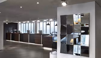 Moderne hotellresepsjonsområde med en rad med innsjekkingsdisker i mørkt tre, hver med en datamaskin. Rommet har elegant belysning og et moderne, reflekterende veggkunstverk. Store vinduer slipper naturlig lys inn og skaper en innbydende atmosfære.