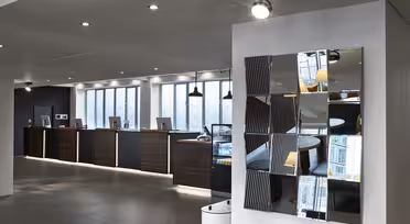 Moderne hotellresepsjonsområde med en rad med innsjekkingsdisker i mørkt tre, hver med en datamaskin. Rommet har elegant belysning og et moderne, reflekterende veggkunstverk. Store vinduer slipper naturlig lys inn og skaper en innbydende atmosfære.
