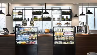 Et moderne kaféinteriør med kjølemontrer fylt med diverse drikkevarer, snacks og pakket mat. Hyllene over inneholder vinflasker og pyntegrønt. Pendellamper henger fra taket, og gir et moderne preg.