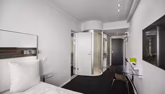 Moderne hotellrom med minimalistisk design. Har en seng med hvitt sengetøy, et bord med glassplate og et åpent, sirkulært dusjområde med frostet glass. Veggene er hvite, og belysningen er subtil, noe som skaper en ren og elegant atmosfære.