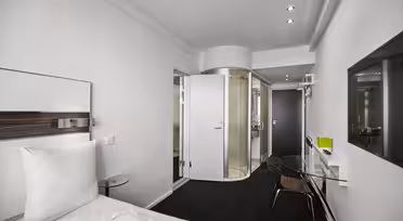 Moderne hotellrom med minimalistisk design. Har en seng med hvitt sengetøy, et bord med glassplate og et åpent, sirkulært dusjområde med frostet glass. Veggene er hvite, og belysningen er subtil, noe som skaper en ren og elegant atmosfære.