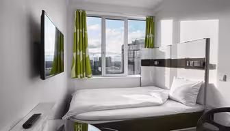 Et moderne, kompakt hotellrom har en enkeltseng med hvitt sengetøy ved siden av et stort vindu med grønne og hvite gardiner. En flatskjerm-TV er montert på veggen. Et skrivebord og en stol med glassplate er i forgrunnen. Bybilde synlig utenfor.