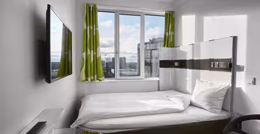 Et moderne, kompakt hotellrom har en enkeltseng med hvitt sengetøy ved siden av et stort vindu med grønne og hvite gardiner. En flatskjerm-TV er montert på veggen. Et skrivebord og en stol med glassplate er i forgrunnen. Bybilde synlig utenfor.