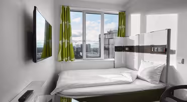 Et moderne, kompakt hotellrom har en enkeltseng med hvitt sengetøy ved siden av et stort vindu med grønne og hvite gardiner. En flatskjerm-TV er montert på veggen. Et skrivebord og en stol med glassplate er i forgrunnen. Bybilde synlig utenfor.