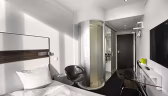 Minimalistisk hotellrom med enkeltseng, hvite sengetøy og hodegjerde. En elegant svart stol sitter ved siden av et sylindrisk dusjkabinett i glass. En veggmontert TV og et glasspult er på motsatt side av sengen. Rommet har en moderne, kompakt design.