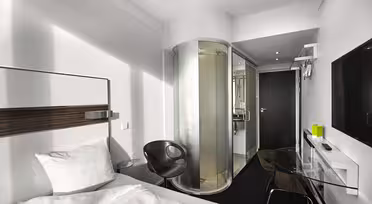 Minimalistisk hotellrom med enkeltseng, hvite sengetøy og hodegjerde. En elegant svart stol sitter ved siden av et sylindrisk dusjkabinett i glass. En veggmontert TV og et glasspult er på motsatt side av sengen. Rommet har en moderne, kompakt design.