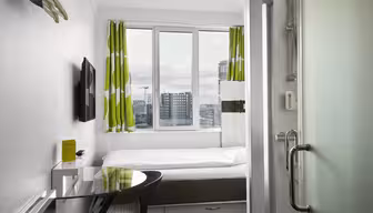 Et kompakt, moderne hotellrom med en enkeltseng under et stort vindu med grønne og hvite gardiner. Rommet har en veggmontert TV, et lite skrivebord med stol og en glassdør som fører til badet. Bybygninger er synlige utenfor.