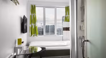 Et kompakt, moderne hotellrom med en enkeltseng under et stort vindu med grønne og hvite gardiner. Rommet har en veggmontert TV, et lite skrivebord med stol og en glassdør som fører til badet. Bybygninger er synlige utenfor.