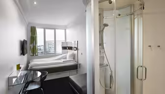Lite moderne hotellrom med en seng nær et vindu, en veggmontert TV og et elegant glasspult med stol. Det er et kompakt, buet dusjkabinett i glass nær inngangen. Grønne gardiner legger til en fargetone. Bybilde synlig gjennom vinduet.