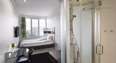 Lite moderne hotellrom med en seng nær et vindu, en veggmontert TV og et elegant glasspult med stol. Det er et kompakt, buet dusjkabinett i glass nær inngangen. Grønne gardiner legger til en fargetone. Bybilde synlig gjennom vinduet.