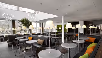 Moderne kaféinteriør med runde bord og polstrede stoler. Hvert bord har en lampe, og sittegruppen har mørkt trekk med grønne og oransje aksentputer. Store vinduer og potteplanter bidrar til den lyse og luftige atmosfæren.