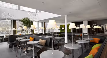 Moderne kaféinteriør med runde bord og polstrede stoler. Hvert bord har en lampe, og sittegruppen har mørkt trekk med grønne og oransje aksentputer. Store vinduer og potteplanter bidrar til den lyse og luftige atmosfæren.