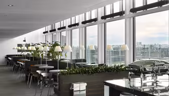 En moderne, romslig kafeteria med store vinduer med utsikt over byen. Den har bord, stoler, innendørs planter og matvarmere i bufféstil. Overheadlys og et elegant design skaper en innbydende atmosfære.