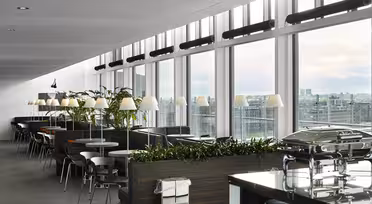 En moderne, romslig kafeteria med store vinduer med utsikt over byen. Den har bord, stoler, innendørs planter og matvarmere i bufféstil. Overheadlys og et elegant design skaper en innbydende atmosfære.