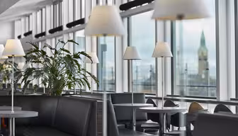 Moderne salong med grå sofaer, runde bord og hvite lamper. Store vinduer avslører et bybilde med et klokketårn i bakgrunnen. Potteplanter gir grønt til den elegante interiørdekorasjonen.