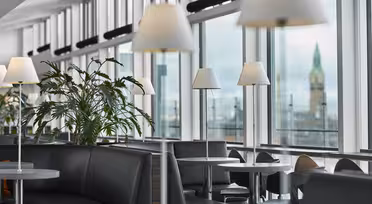 Moderne salong med grå sofaer, runde bord og hvite lamper. Store vinduer avslører et bybilde med et klokketårn i bakgrunnen. Potteplanter gir grønt til den elegante interiørdekorasjonen.