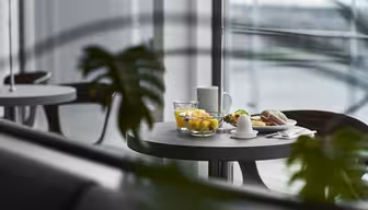 Et rundt bord med frokostartikler, inkludert en skål med frukt, et glass vann, en kaffekopp, en smørskål og et bakverk, sett gjennom grønne planteblader i en moderne spisestue med store vinduer.